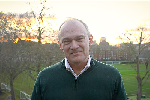 Ed Davey. Ed is wearing a green jumper over a shirt. In the phot background are trees, an open green space, buildings and the setting sun.